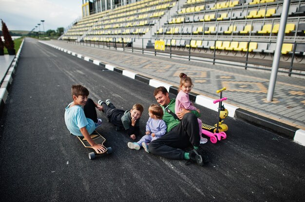 Quattro bambini con il padre in asfalto giocano e si divertono. La famiglia sportiva trascorre il tempo libero all'aperto con scooter e pattini.