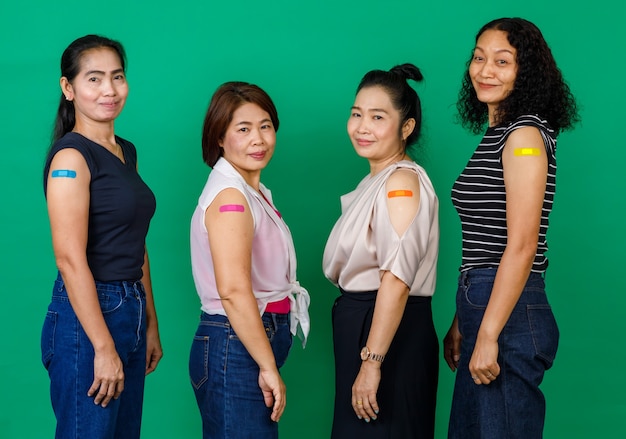 Quattro donne asiatiche di mezza età che mostrano le braccia con una benda che mostra che sono state vaccinate per il virus Covid 19 su sfondo verde. Concetto per la vaccinazione contro il Covid 19.
