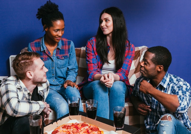 Quattro giovani amici interrazziali si siedono sul divano di casa con una pizza fresca