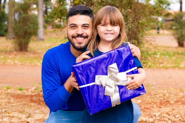 Questo regalo è per te Ritratto di una bambina amorevole accanto al padre che lo saluta con la festa del papà o il compleanno che tiene una confezione regalo incartata felice vacanza in famiglia insieme nel parco