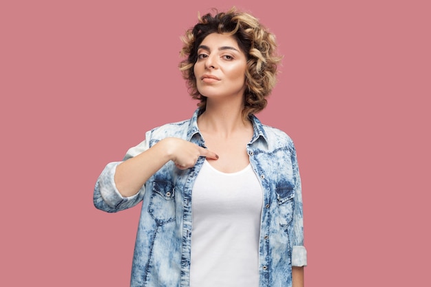 Questo sono io. Ritratto di giovane donna seria con l'acconciatura riccia in camicia blu casual in piedi, indicandosi con il viso orgoglioso e guardando la fotocamera. girato in studio al coperto, isolato su sfondo rosa.