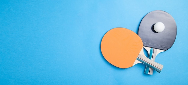 Racchette da ping pong e una pallina di plastica bianca su sfondo blu.