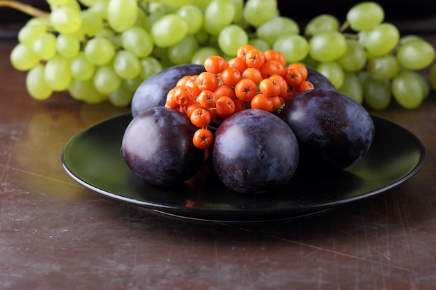 Raccolta di frutti autunnali crudi e biologici dal primo piano del giardino Uva succosa matura prugna e sorbo su una piastra nera su sfondo scuro