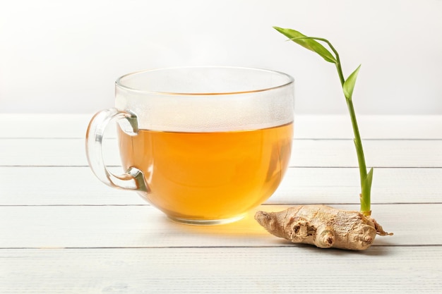 Radice di zenzero secca con germoglio verde su tavola di cinghiale bianco, tazza di vetro di tè appena fatto accanto ad essa.