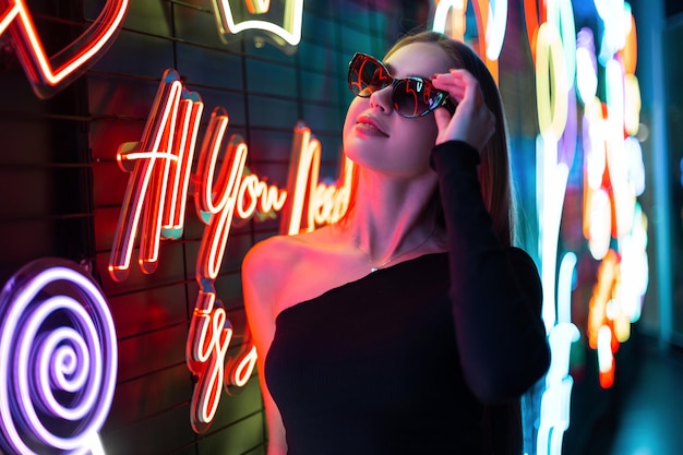 Raffinata modella giovane donna alla moda in abiti neri di moda indossa occhiali da sole e cammina vicino alle luci al neon colorate dei segni