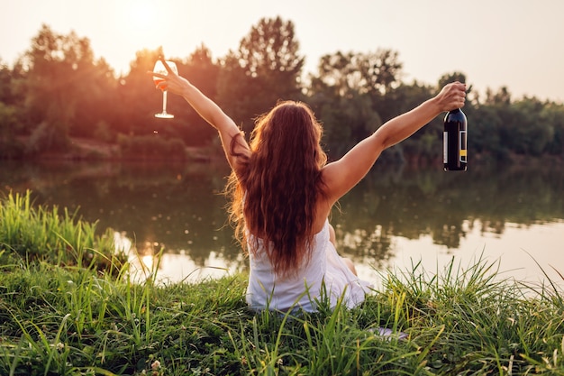 Raffreddare con il vino. Donna che gode del bicchiere di vino sulla sponda del fiume al tramonto che alza le armi e che si sente libero e felice.