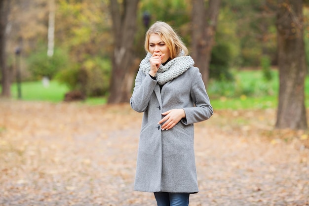 Raffreddore e influenza. Giovane donna in un cappotto grigio che cammina nel parco autunnale e riscalda la mano congelata.