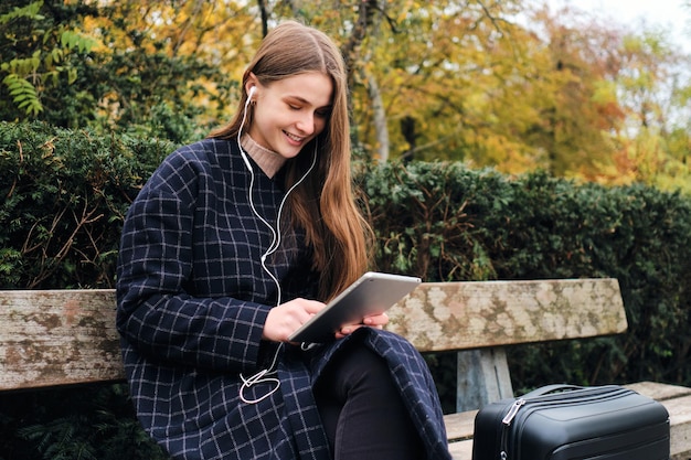 Ragazza abbastanza casual in auricolari felicemente utilizzando tablet seduto con la valigia sulla panchina nel parco cittadino