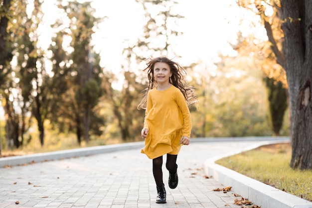 Ragazza abbastanza divertente per bambini 56 anni, vestito giallo elegante, correre nel parco autunnale con foglie cadute all'aperto Felicità dell'infanzia