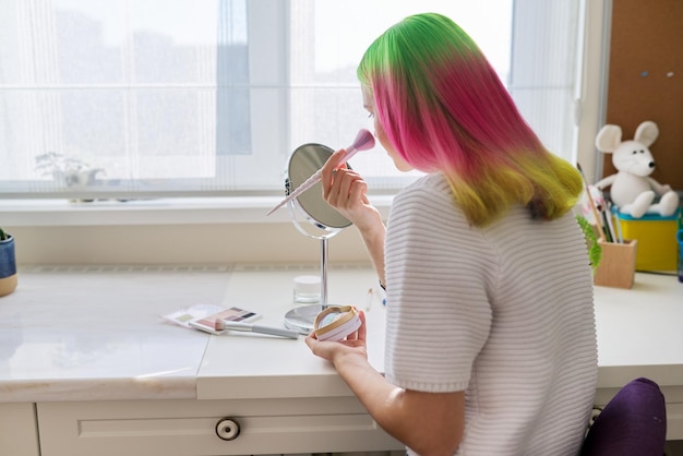 Ragazza adolescente alla moda alla moda con capelli colorati tinti che fa il trucco guardandosi allo specchio con una spazzola di cosmetici decorativi seduti a tavola a casa