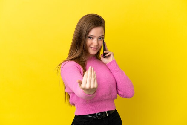 Ragazza adolescente che utilizza il telefono cellulare su sfondo giallo isolato che invita a venire con la mano. Felice che tu sia venuto