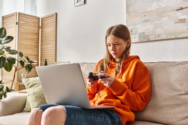 Ragazza adolescente con joystick e laptop che gioca e si siede sul divano a casa.