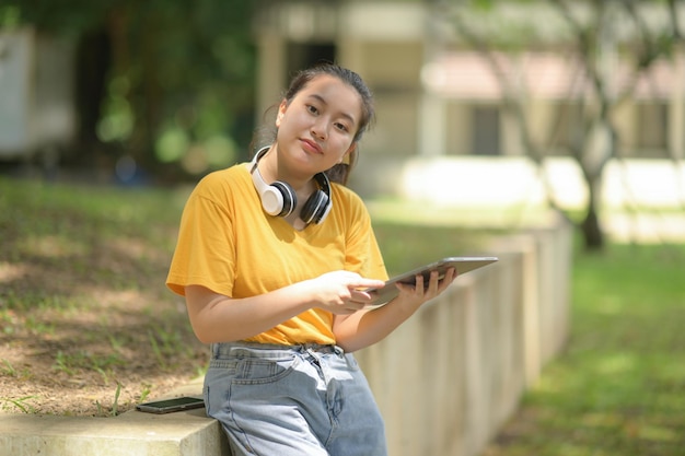 Ragazza adolescente con la camicia gialla che indossa le cuffie sul collo, con in mano un tablet in piedi in giardino guardando la telecamera, lavoro da casa, lavoro a distanza, ufficio a casa, studio online.