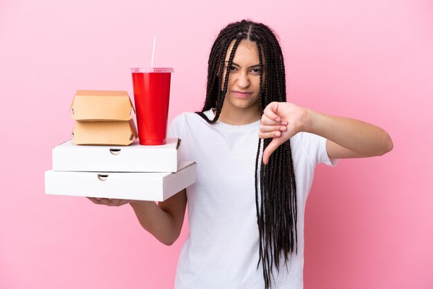 Ragazza adolescente con le trecce che tengono pizze e hamburger su sfondo rosa isolato che mostra pollice giù con espressione negativa