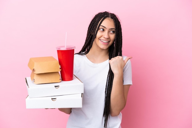 Ragazza adolescente con le trecce che tengono pizze e hamburger su sfondo rosa isolato che punta al lato per presentare un prodotto