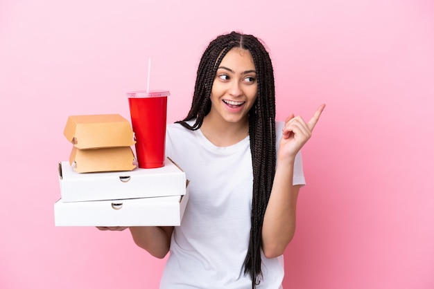 Ragazza adolescente con trecce che tengono pizze e hamburger su sfondo rosa isolato con l'intenzione di realizzare la soluzione mentre si solleva un dito