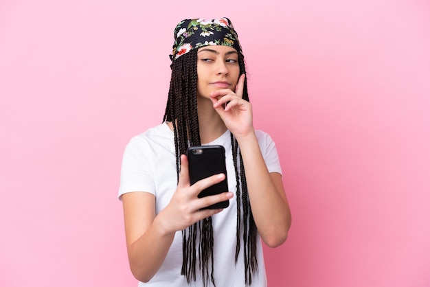 Ragazza adolescente con trecce su sfondo rosa isolato utilizzando il telefono cellulare e il pensiero