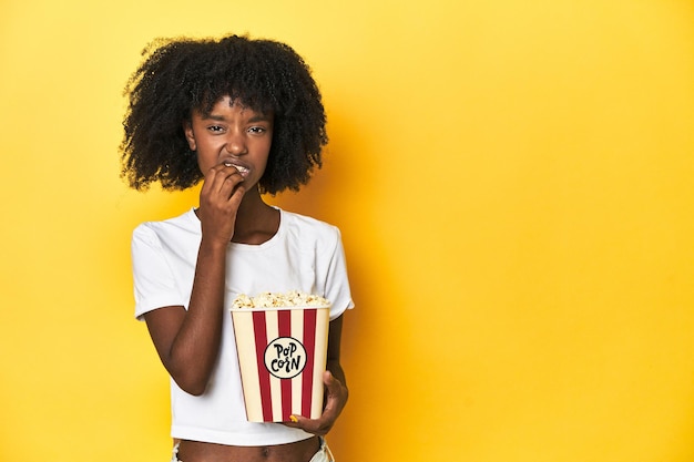 Ragazza adolescente con un concetto di cinema di popcorn su uno sfondo giallo che si morde le unghie nervosa e molto ansiosa