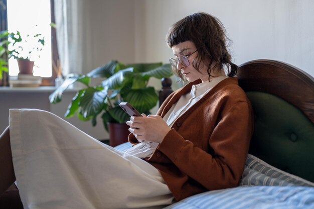 Ragazza adolescente concentrata e pensierosa che usa il telefono cellulare e scorre sui social media mentre si rilassa a casa