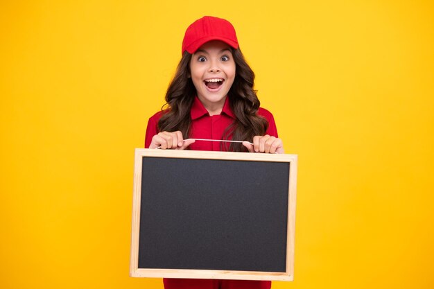 Ragazza adolescente eccitata tenere lavagna Ragazza bambino eccitata in uniforme rossa e cappuccio Ragazza adolescente tenere lavagna isolata su sfondo giallo Copia spazio sul bordo vuoto mock up