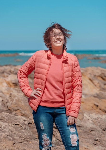 Ragazza adolescente felice che indossa una camicia rosa e una giubba pudded all'aperto vicino al mockup del mare