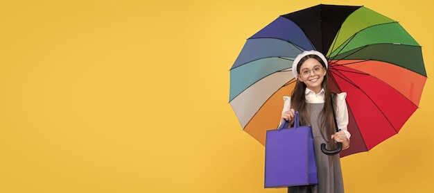Ragazza adolescente felice sotto l'ombrello colorato nella stagione autunnale tenere la borsa della spesa vendita autunnale Bambino con ombrello autunnale tempo piovoso banner poster orizzontale con spazio per la copia