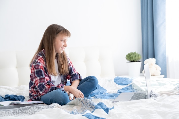 ragazza adolescente guardando i video sul portatile seduto sul letto nella sua stanza. studente adolescente che studia online, apprendimento a distanza,