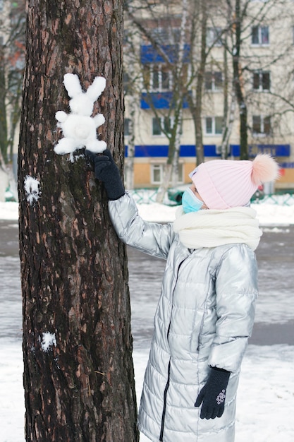 Ragazza adolescente in mascherina medica con lepre fatta a mano o coniglio di neve bianca sulla corteccia di albero