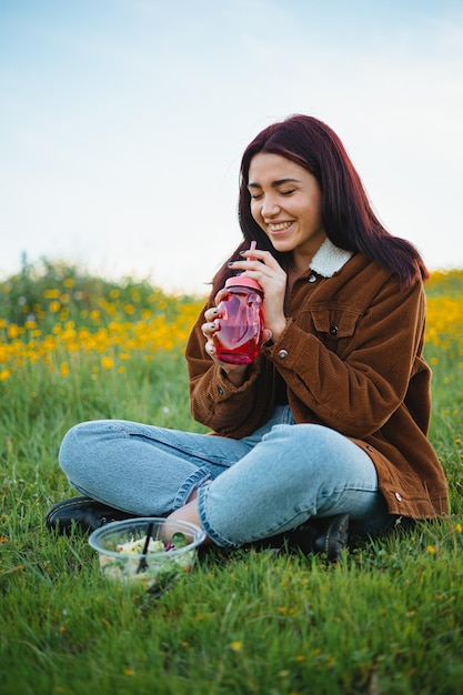 Ragazza adolescente ridente che beve in un barattolo rosso seduto sull'erba di una collina Ha un'insalata accanto a lei