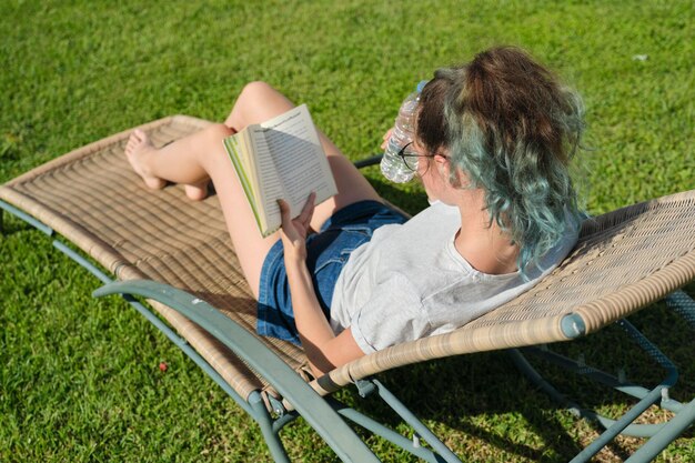 Ragazza adolescente rilassante seduta all'aperto su una sedia in rattan sul prato verde, leggendo un libro e bevendo acqua dalla bottiglia, soleggiata giornata estiva