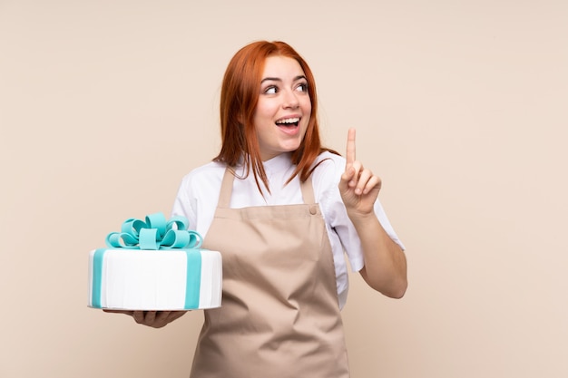 Ragazza adolescente rossa con una grande torta che intende realizzare la soluzione sollevando un dito