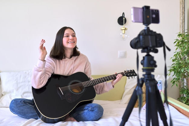 Ragazza adolescente seduta a casa sul letto con una chitarra acustica, ragazza che suona la chitarra registrando video sulla telecamera. Tecnologia, social network, arte, hobby, concetto di adolescenti