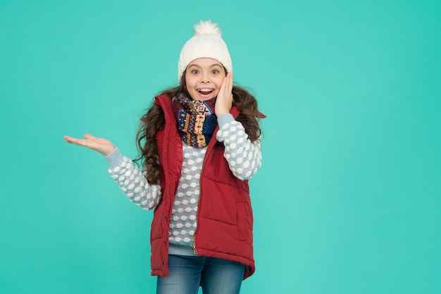 Ragazza adolescente sorpresa con lunghi capelli ricci indossare gilet imbottito e maglieria durante le vacanze di Natale che presenta lo spazio della copia del prodotto vendite invernali