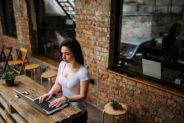 Ragazza adorabile dello studente che utilizza computer portatile e le cuffie mentre stando Muro di mattoni sullo sfondo.