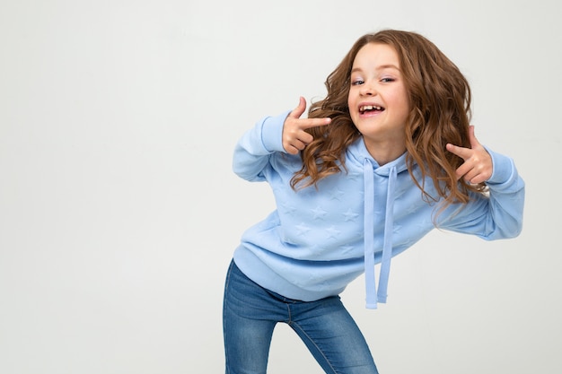 Ragazza affascinante in una maglia con cappuccio blu che posa pendente in avanti su una parete bianca con spazio