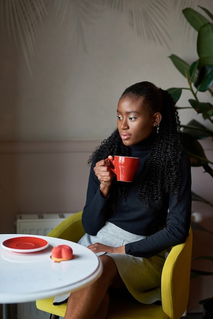 ragazza africana alla moda seduta al tavolino del bar godendosi il cappuccino mattutino