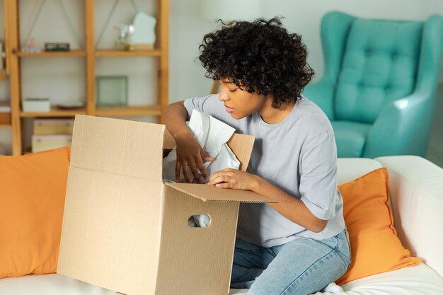 Ragazza africana che disimballa la consegna guardando nella scatola Donna felice che apre la scatola di cartone Femmina che riceve il pacco guardando gli articoli della merce consegnata Feedback positivo del cliente soddisfatto consegna espressa rapida