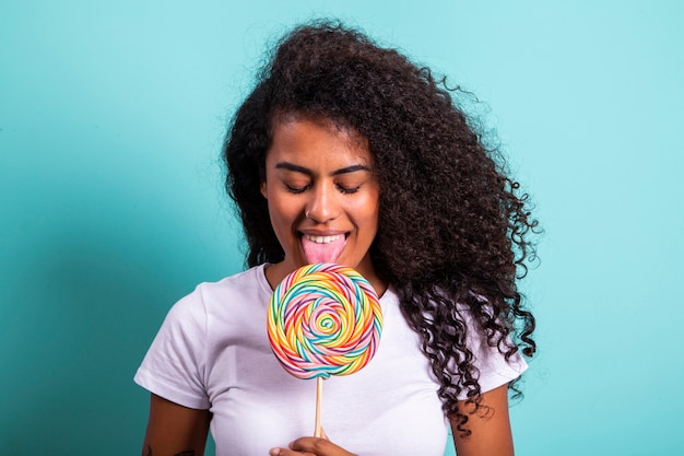 Ragazza africana riccia sorridente in maglietta bianca brillante che tiene il lecca-lecca della caramella. Giovane donna allegra che nasconde la faccia anteriore con il lecca-lecca