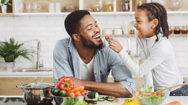 Ragazza africana sveglia che dà al suo papà pomodoro ciliegino durante la cottura