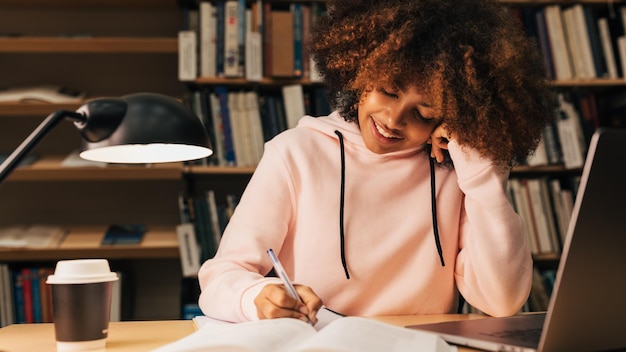 Ragazza afroamericana che legge dal libro in biblioteca mentre prepara gli esami la sera Studente seduto alla scrivaniax9xA