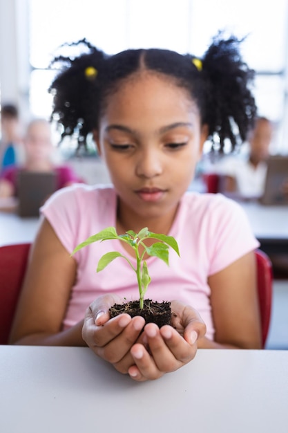 Ragazza afroamericana che tiene una piantina della pianta mentre si siede sulla sua scrivania in classe a scuola