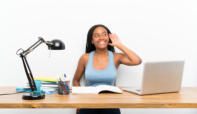 Ragazza afroamericana dello studente dell'adolescente con capelli intrecciati lunghi nel suo posto di lavoro che ascolta qualcosa