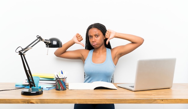 Ragazza afroamericana dello studente dell'adolescente con capelli intrecciati lunghi nel suo posto di lavoro che mostra pollice giù