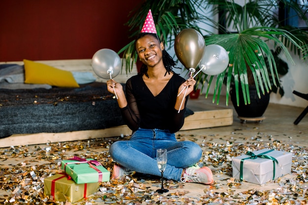 Ragazza afroamericana divertente in cappello di compleanno e con gli aerostati che si siedono sul pavimento con i regali e i coriandoli delle scatole attuali. Celebrazione, concetto di festa