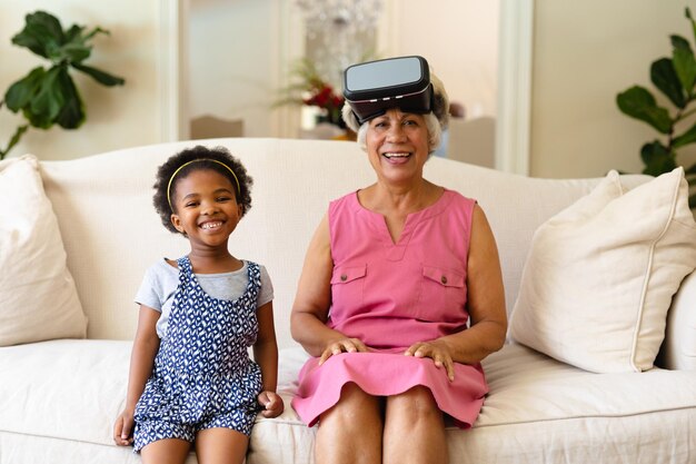 Ragazza afroamericana e sua nonna che indossano cuffie vr sorridenti mentre si siedono sul divano