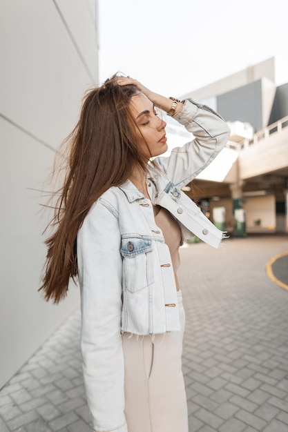 Ragazza alla moda carina e pacifica in abiti di jeans alla moda che camminano per la città e si aggiustano i capelli