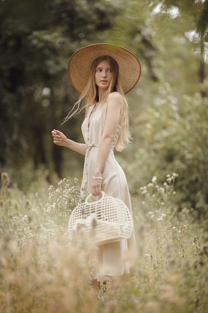 Ragazza alla moda che cammina attraverso il prato con un cesto di fiori di campo. Giovane donna bionda naturale