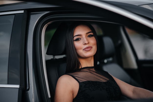 Ragazza alla moda che si siede in un'automobile del Business class in un vestito nero. Moda e stile aziendale