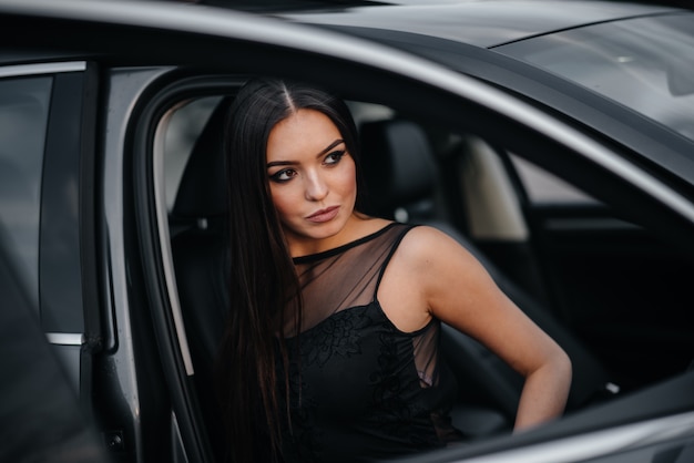 Ragazza alla moda che si siede in un'automobile del Business class in un vestito nero. Moda e stile aziendale