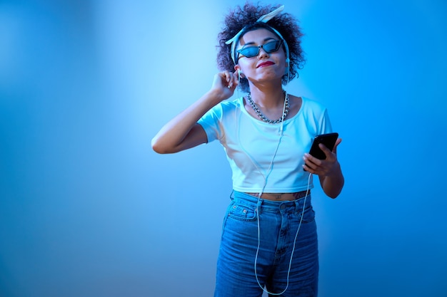 Ragazza alla moda con riccioli afro ascolta e gode di musica con le cuffie in luce al neon blu, balli modello stile hip hop e relax